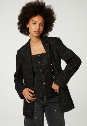 Schwarzer strukturiert Blazer mit taillierter Passform, ausgestattet mit einem einzelnen Knopfverschluss und goldenen Hardware-Details. Wird über einem passenden Oberteil getragen.