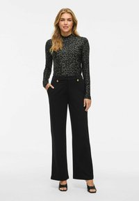 Vrouw die staat en een lang mouwloze top met luipaardprint draagt, hoge taille zwarte wijde broek met gouden knopen, en zwarte open teenhakken.