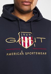 Felpa blu navy con cappuccio e cordoncino, presenta uno scudetto ricamato in oro e rosso con la scritta "GANT AMERICAN SPORTSWEAR". Materiale in morbido cotone misto.