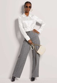 Vrouw in een witte satijnen blouse met knopen, grijze wijde broek, zwarte laarzen, grote oorbellen, ronde zonnebril en een crèmekleurige handtas.