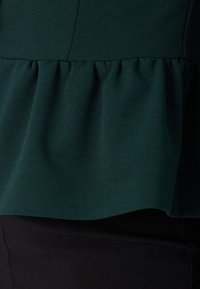 Dark green peplum top with soft, textured fabric; features subtle gathers along the hem and pairs with a black bottom.