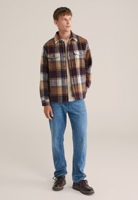 Bunda s fleece podšívkou, károvaná v burgundskej, béžovej a modrej farbe s dvoma prednými vreckami. Nosená cez svetlú košeľu a skombinovaná s modrými džinami a tmavými topánkami.