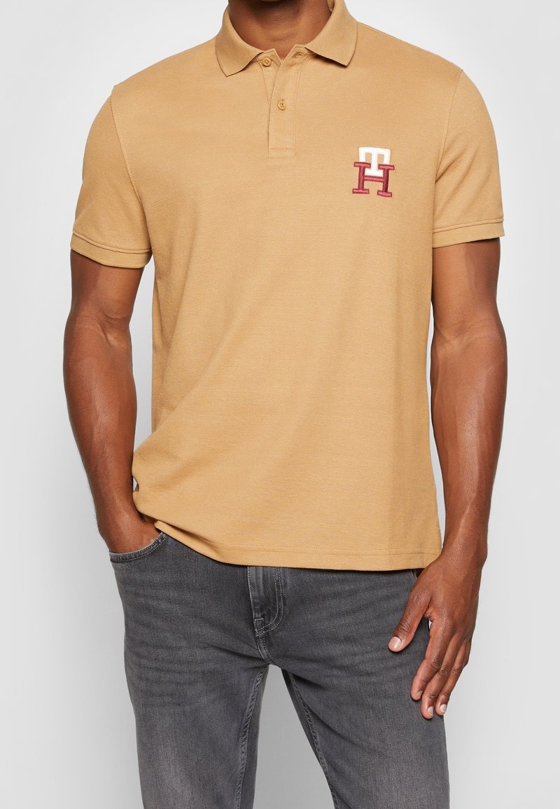 Polo à manches courtes en coton beige clair, présentant une finition texturée et un logo bordeaux et blanc sur la poitrine gauche. Associé à un jean gris foncé.