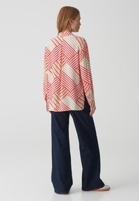 Camicia a maniche lunghe con un motivo a righe diagonali rosse e crema, realizzata in un tessuto leggero, indossata con pantaloni larghi blu scuro.