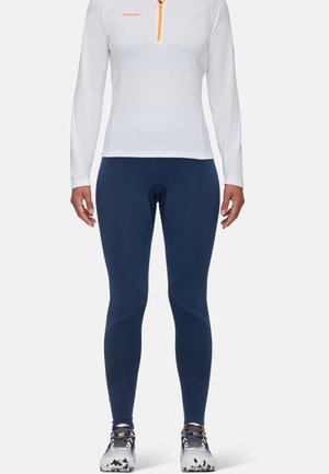Top blanc à manches longues avec fermeture éclair orange et logo, associé à un legging marine ajusté avec des accents texturés et des détails réfléchissants.