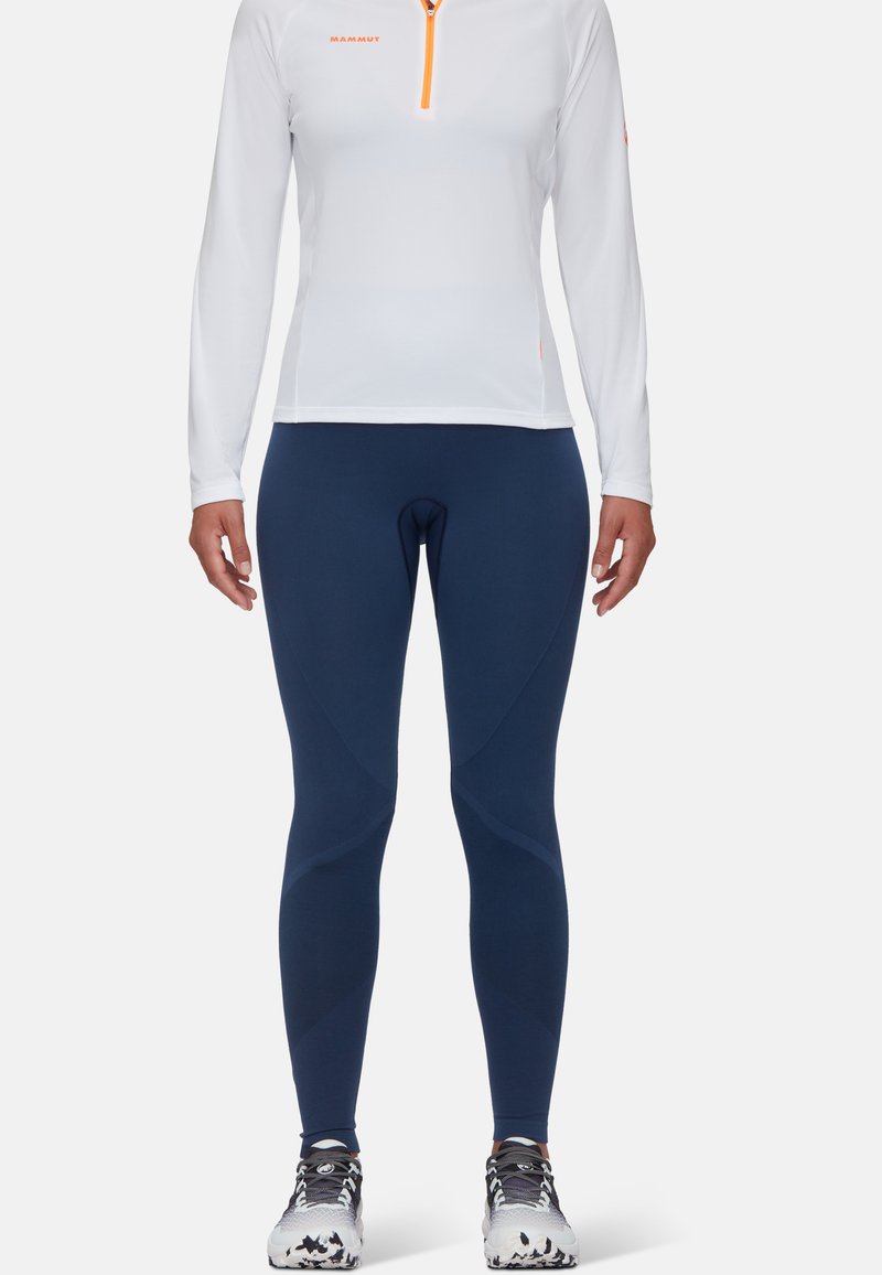 Maglia bianca a maniche lunghe con zip arancione e logo, abbinata a leggings attillati blu navy con dettagli testurizzati e riflettenti.