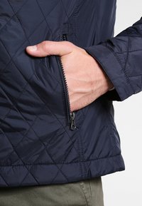 Marinblå vadderad jacka med en dragkedjeficka, som har en slät yta och en ställbar krage. Syntes mönster av sömmarna längs tyget.