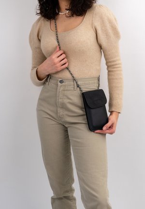 Femme portant un pull beige et un pantalon, tenant un petit sac bandoulière noir à chaîne, sur un fond uni.