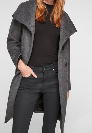 Vrouw met rood haar in een donkergrijze wollen jas over een zwart getailleerd top en zwarte broek, met een hand in haar zak, tegen een effen achtergrond.