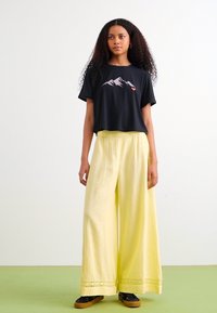Zwarte cropped t-shirt met bergafbeelding, gecombineerd met vloeiende, hooggetailleerde gele wijde broek met kantafwerking. Zwarte schoenen maken de look af.