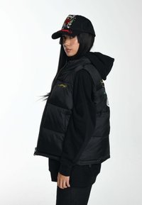 Schwarze gepolsterte Weste mit gelber Logo-Stickerei, getragen über einem schwarzen Hoodie. Eine schwarze Kappe mit bunten Grafiken vervollständigt das Outfit.