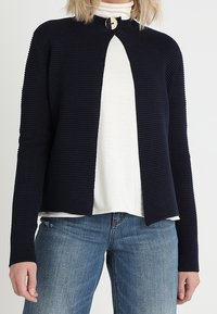 Námořnický žebrovaný kardigan s vysokým límcem, otevřeným předním dílem a kulatým detailem knoflíku. Nosený přes bílý rolák a modré džíny.