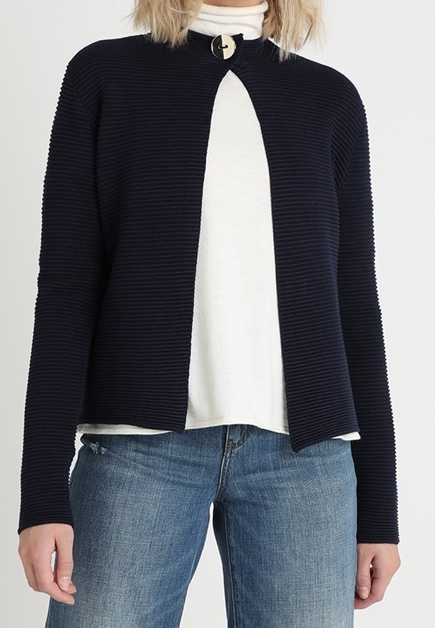 Námořnický žebrovaný kardigan s vysokým límcem, otevřeným předním dílem a kulatým detailem knoflíku. Nosený přes bílý rolák a modré džíny.