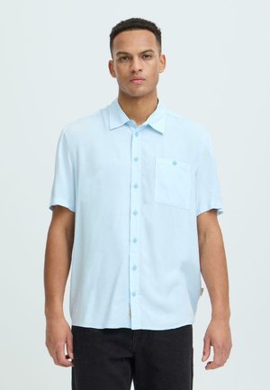 Homme debout portant une chemise à manches courtes bleu clair avec une poche poitrine et un pantalon noir, sur un fond uni.