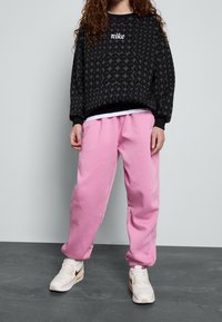 Černá mikina se vzorem a bílým logem "Nike", doplněná volnými růžovými tepláky a béžovými teniskami s burgundskými akcenty.
