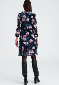 Robe à volants florale en bleu marine foncé, ornée de grandes roses aux nuances de rose et de bleu-vert. Manches longues et taille élastiquée, assortie à des bottes noires montantes jusqu'aux genoux.