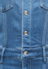 Τζάκετ από μπλε τζιν ύφασμα μπροστά με ασημένια μεταλλικά κουμπιά, ορατές ραφές και δύο τσέπες στο στήθος στις πάνω άκρες.