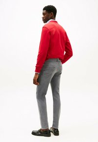 Czerwona bluza, szare obcisłe dżinsy, czarne loafersy. Model stoi tyłem do kamery, z widocznymi ryflowanymi mankietami i kołnierzykiem pod spodem.