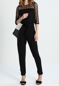Black jumpsuit with a sheer, dotted mesh top and long sleeves. Features a drawstring waist and tapered legs. Worn with a small silver handbag.
