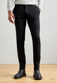 Pantalones negros ajustados de tela suave, con bolsillos laterales y frente plano. Combinados con botas negras de tobillo sobre un suelo de madera.