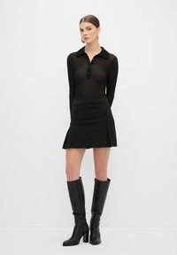 Top negro de manga larga con patrón de malla y cuello, combinado con una falda corta negra con pliegues, complementado con botas negras hasta la rodilla.