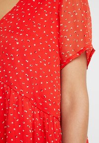 Robe rouge avec manches courtes et transparentes. Présente un motif à pois blancs et noirs. Design fluide avec des fronces à la taille.