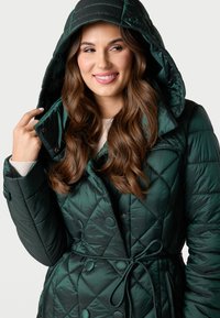 Zelený prošívaný puffer kabát s kapucí, který má velké knoflíky a pásový pas. Tkanina má hladkou, lesklou strukturu.