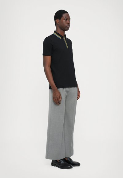 Polo negro con un cuello a rayas amarillas, combinado con pantalones grises anchos y zapatos negros, mostrado sobre un fondo blanco.
