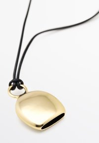 Pendente dorato su una collana di corda nera, caratterizzato da una forma liscia e arrotondata con un lato aperto e un attacco a anello metallico circolare.