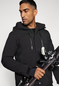 Schwarze Skijacke mit Kapuze, zwei Reißverschluss-Brusttaschen und strukturierter Oberfläche. Die Person hält einen Ski mit Bindungen und einen Skistock.
