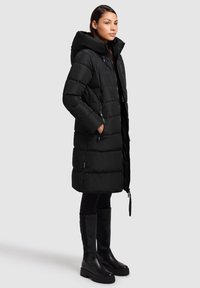 Zwarte pufferjas met capuchon, voorzien van een doorgestikt patroon, ritssluiting aan de voorkant en zijzakken. Draagt bijpassende zwarte legging en kniehoge laarzen.