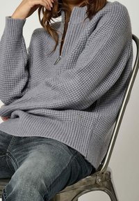 Pull en maille gris clair avec col à fermeture éclair partielle, motif texturé, poignets et ourlet côtelés. Associé à un jean en denim foncé.