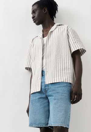 Dospelý s tmavou pokožkou, oblečený v béžovej a bielej vertikálne pruhovanej krátkovej košeli, bielom tielku a svetlomodrých džínsových kraťasoch, stojaci pri bielej stene.