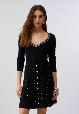 Ženska oblečena v črno obleko z V-izrezom in dolgimi rokavi, okrašeno s srebrnimi zatiči na izrezu in krilu, dopolnjena s srebrnimi uhani in zapestnicami.