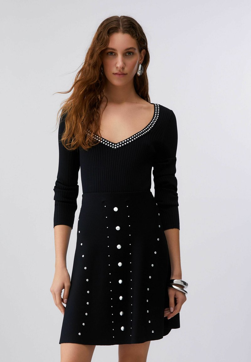 Femme portant une robe noire à manches longues avec un décolleté en V, ornée de clous argentés sur l'encolure et la jupe, accessoirisée avec des boucles d'oreilles et des bracelets argentés.