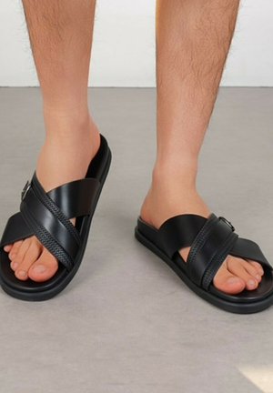 Pieds portant des sandales slides noires avec de larges brides superposées et un détail texturé sur un sol gris.