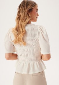 Blusa peplum bianca con smock e maniche corte a puff in tessuto testurizzato. La parte posteriore presenta dettagli arricciati per una silhouette aderente.