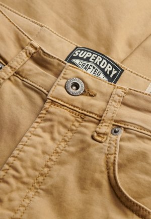 Acercamiento de pantalones beige cosidos con botón de metal y etiqueta de la marca "Superdry Crafted" en blanco y negro visible.