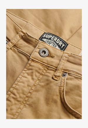 Close-up of tan stitched pants with metal button and visible black and white "Superdry Crafted" brand label.
