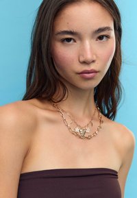 Guld-tonet lagdelt halskæde med hjerte- og blomstercharms, med forlængede ovale led. Modellen bærer en brun stropløs top mod en blå baggrund.