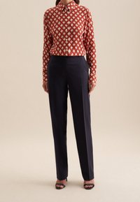 Kvinna som bär en långärmad blus med röd och vit geometrisk mönstring, marinblå skräddarsydda byxor och svarta öppna klackskor stående framför en beige bakgrund.