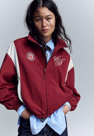 Mladá žena s dlhými tmavými vlasmi, oblečená v červeno-bielej bunde na zips cez svetlomodrú košeľu, stojaca pred jednoduchým pozadím.