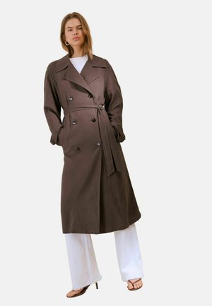 Donna che indossa un lungo trench coat marrone con cintura sopra pantaloni bianchi e sandali neri con tacco alto, in piedi con le mani nelle tasche.