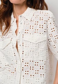 Witte katoenen shirt met uitsnijdingen, korte mouwen, knopen aan de voorkant en twee borstzakken. Gestructureerd patroon met cirkelvormige gaten.