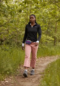 Zwarte fleece-jas met ritssluiting, lichtroze cropped broek, multicolore heuptas, grijze wandelschoenen. Natuurlijke achtergrond met groene bladeren.