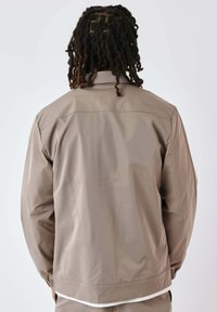 Giacca leggera beige con una superficie liscia, polsini elasticizzati e dettaglio di cucitura sul retro. Senza logo o decorazioni visibili.