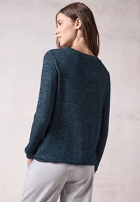 Tumman teal-värinen neulepaita pitkillä hihoilla ja leveällä kaula-aukolla, jossa on teksturoitu neulos. Yhdistetty vaaleanharmaisiin housuihin, mikä luo rennon ilmeen.