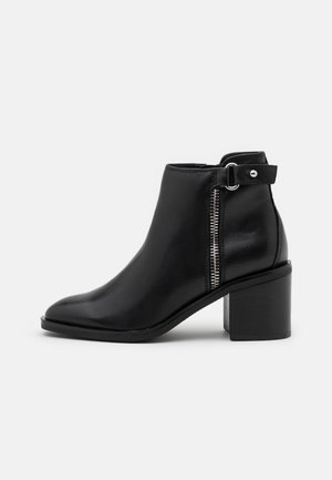 Bottine en cuir noir avec talon carré, fermeture éclair latérale et bride réglable avec détail boucle.