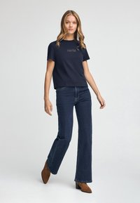 Camiseta de algodón azul marino con mangas cortas, decorada con bordado marrón claro. Combinada con pantalones de mezclilla oscuros acampanados y botines marrones.