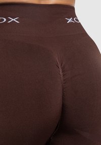 Leggings marroni a vita alta realizzati in materiale elastico, con una texture liscia, dettagli di cucitura arricciata e logo bianco in vita.